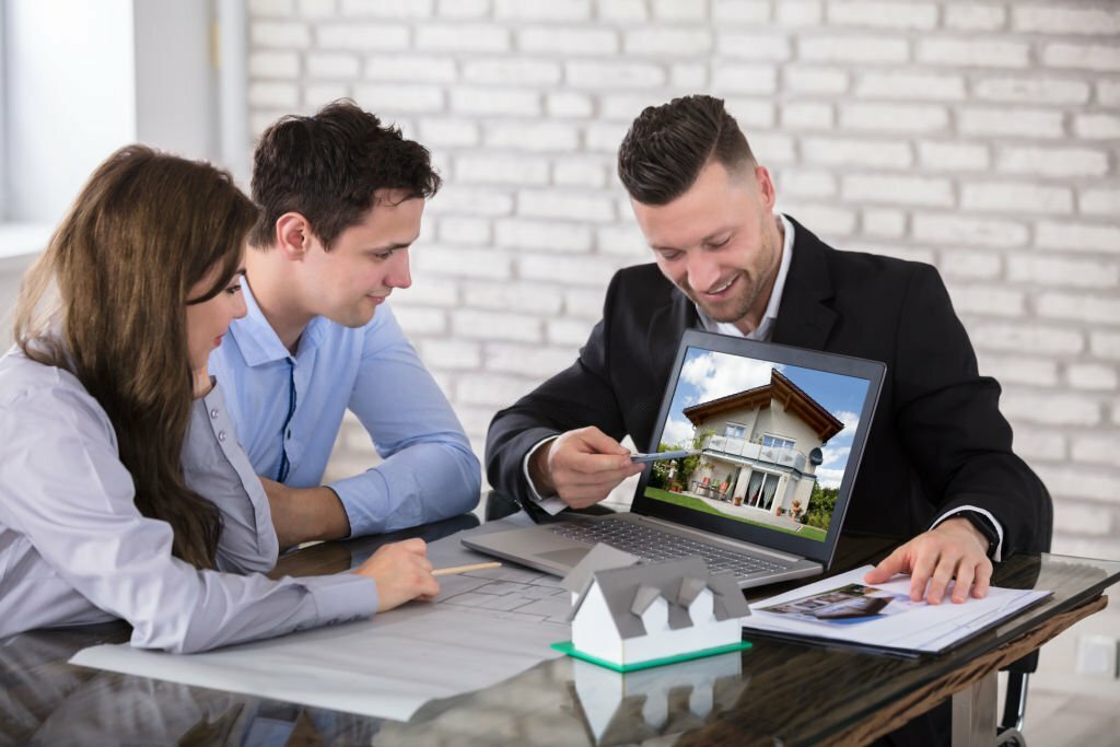 Male Architect Showing House Model On Laptop To Couple In Office